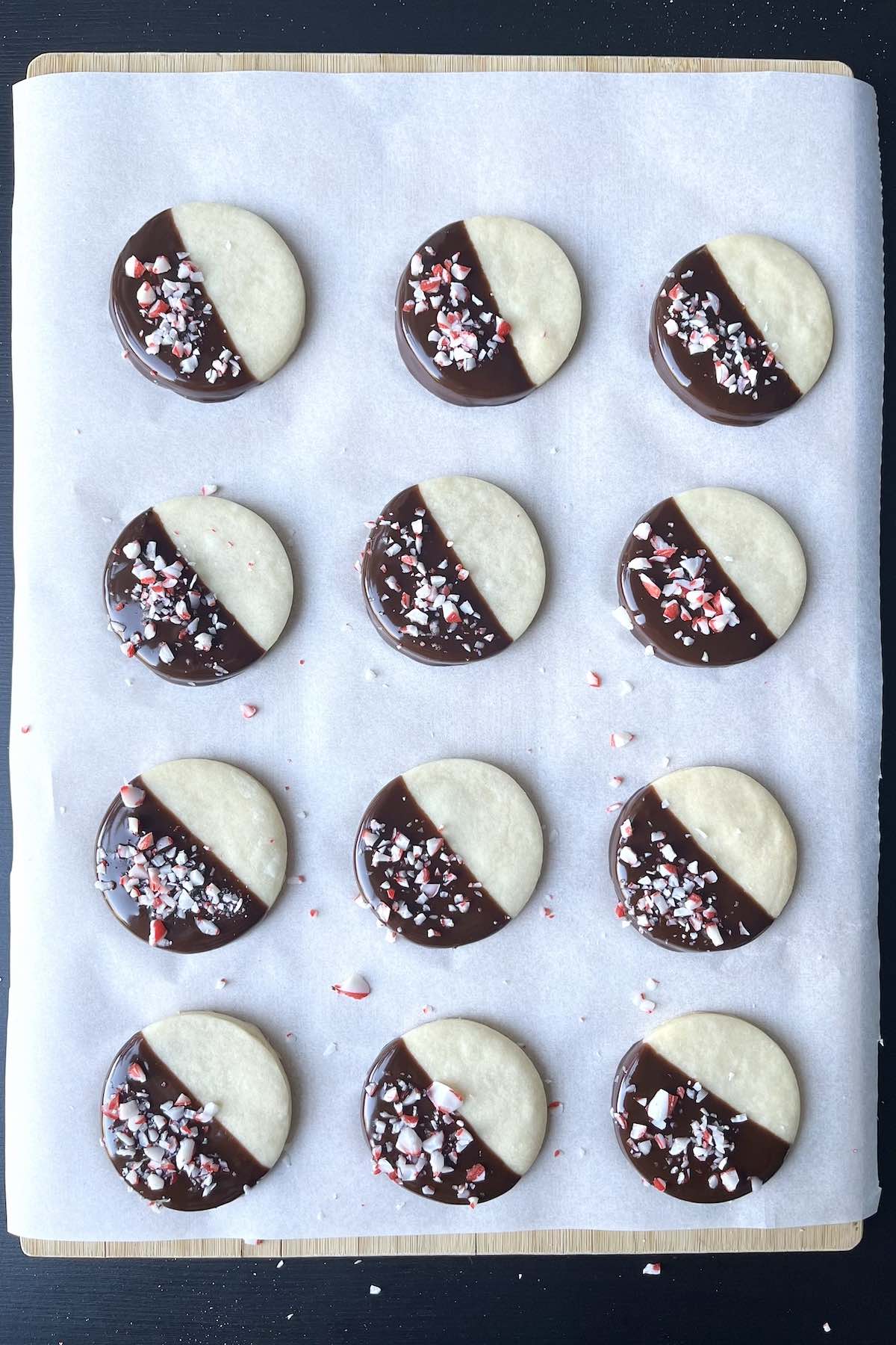 Shortbread cookies dipped in chocolate and sprinkles with crushed candy cane scattered on a piece of parchment paper. 