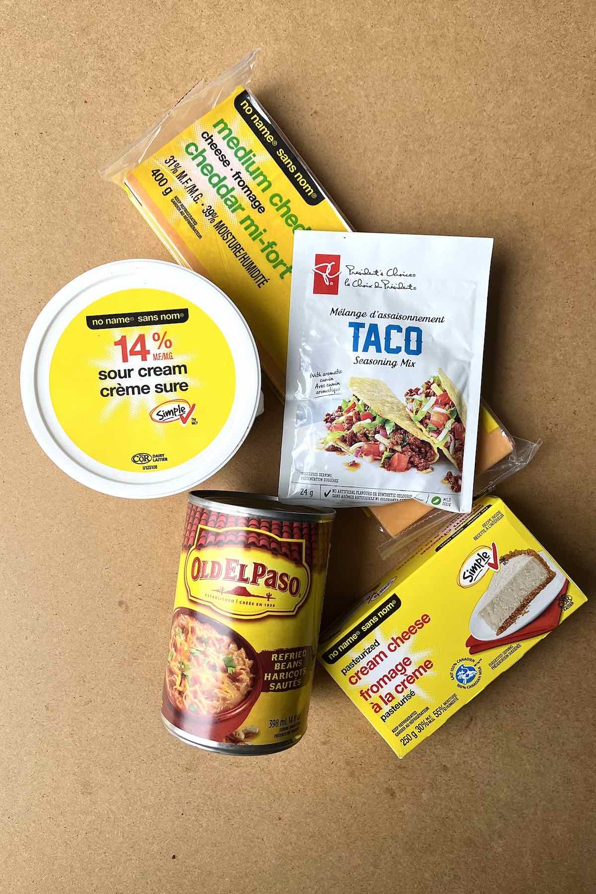 Ingredients needed to make the dip including sour cream, cream cheese, cheddar cheese, taco seasoning, and canned refried beans.