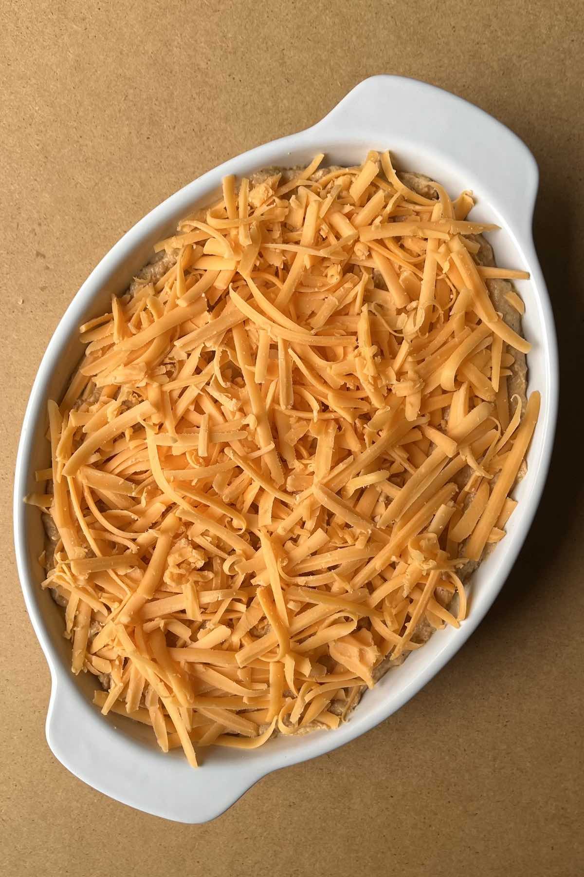 Unbaked refried bean dip in a white baking dish topped with shredded cheddar cheese.