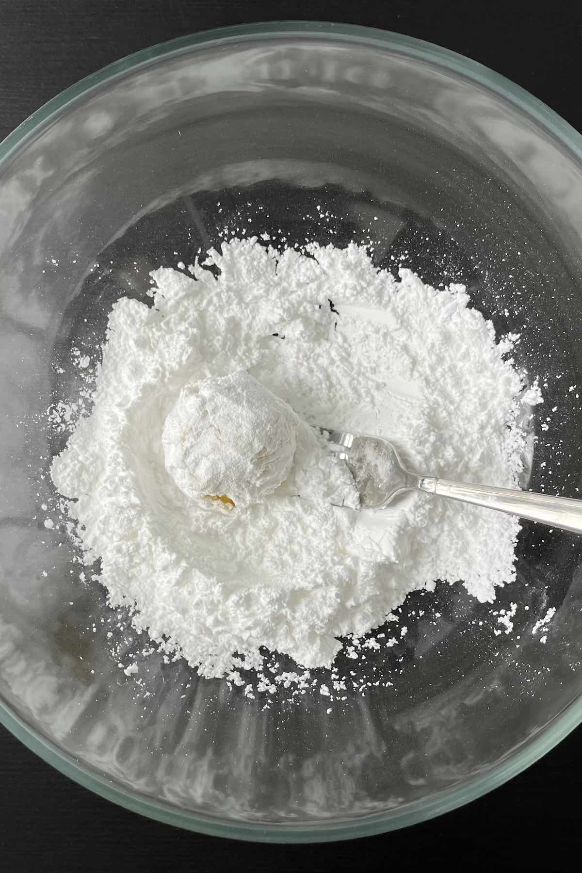 Cookie dough ball tossed in a bowl filled with powdered sugar.