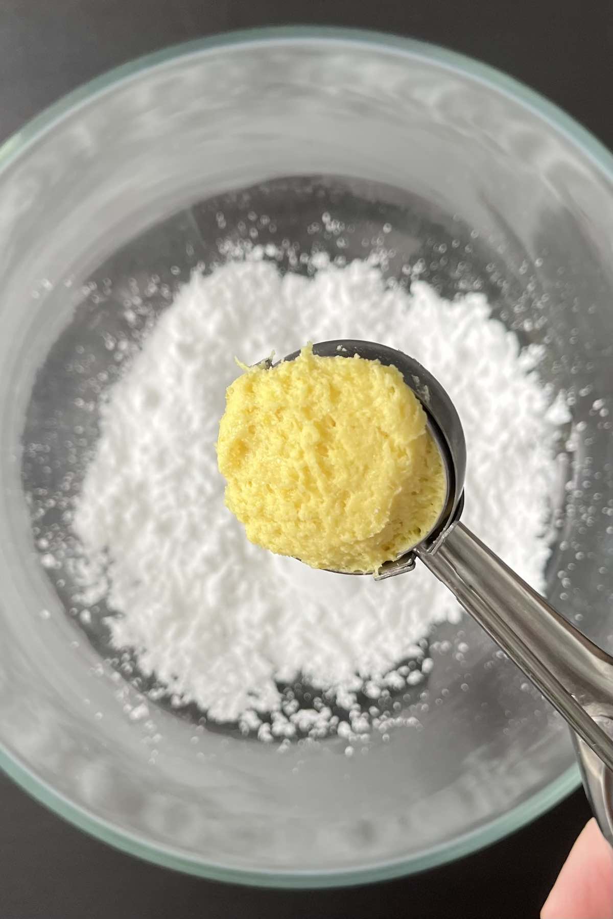 Cookie scoop holding a scoop of cookie dough above a bowl filled with powdered sugar.