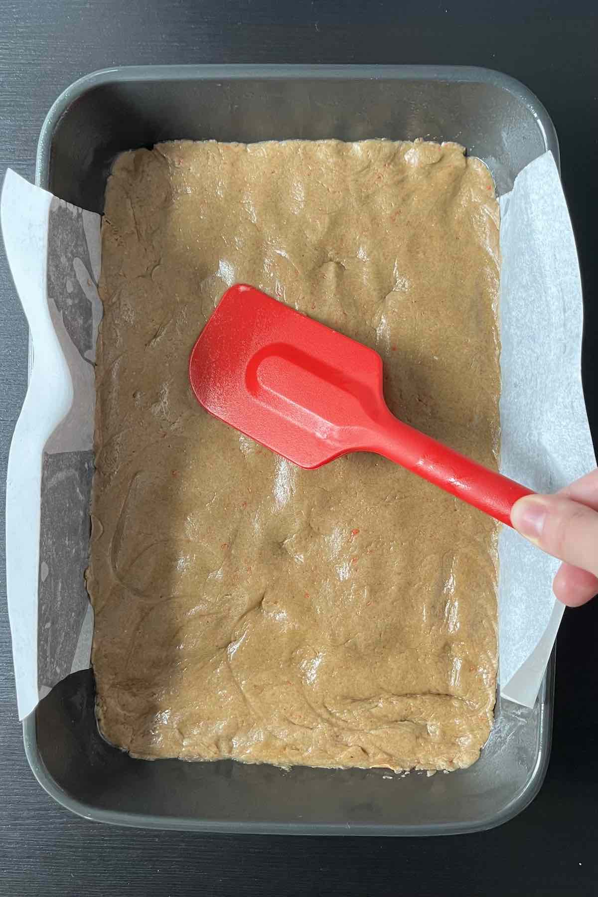 Carrot cake bar batter being pressed into a lined baking dish by a rubber spatula. 