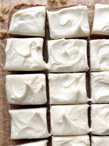 Carrot cake bars with cake mix scattered on a brown piece of parchment paper with chopped walnuts around them.
