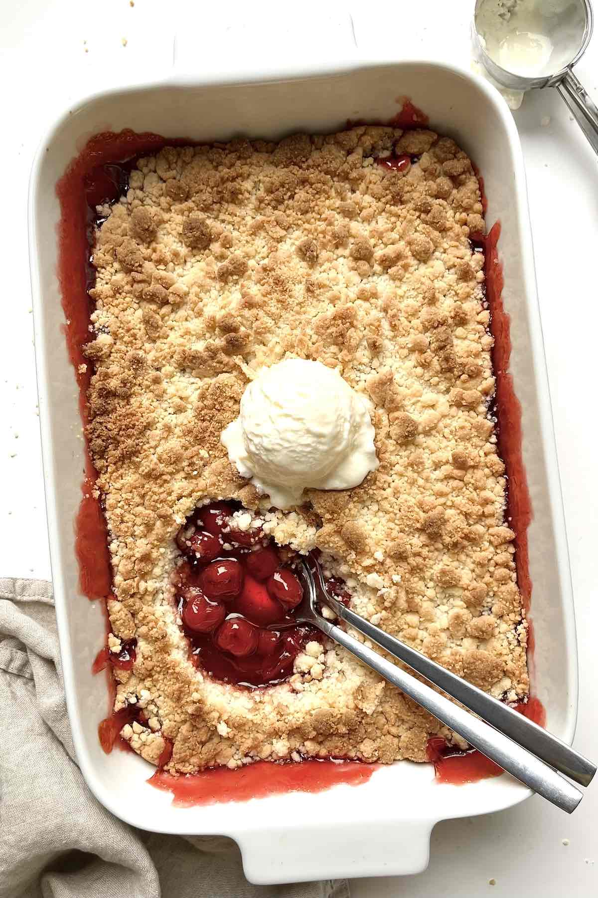 Cherry cobbler with cake mix in a white baking dish topped with a scoop of vanilla ice cream.