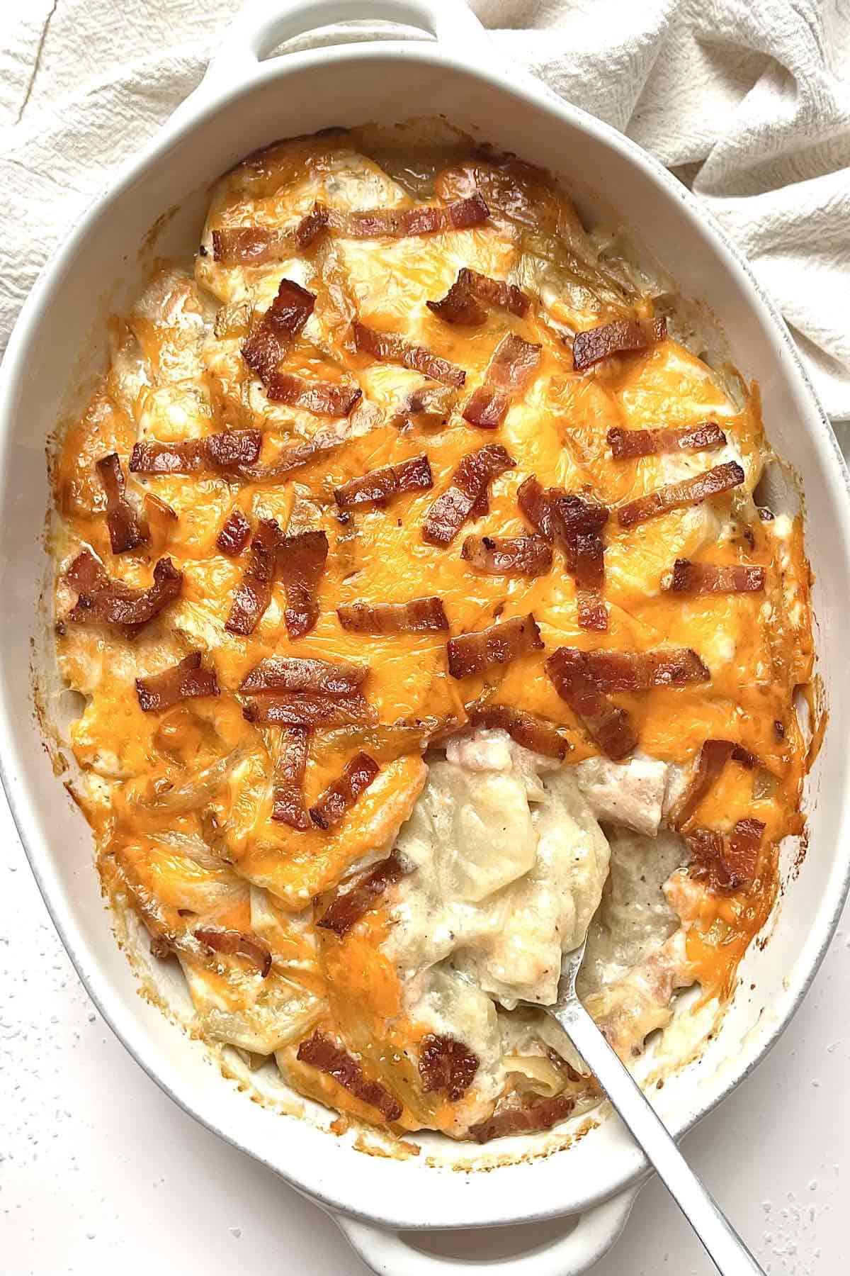 Loaded scalloped potatoes with bacon in a white baking dish with a metal serving spoon in it.