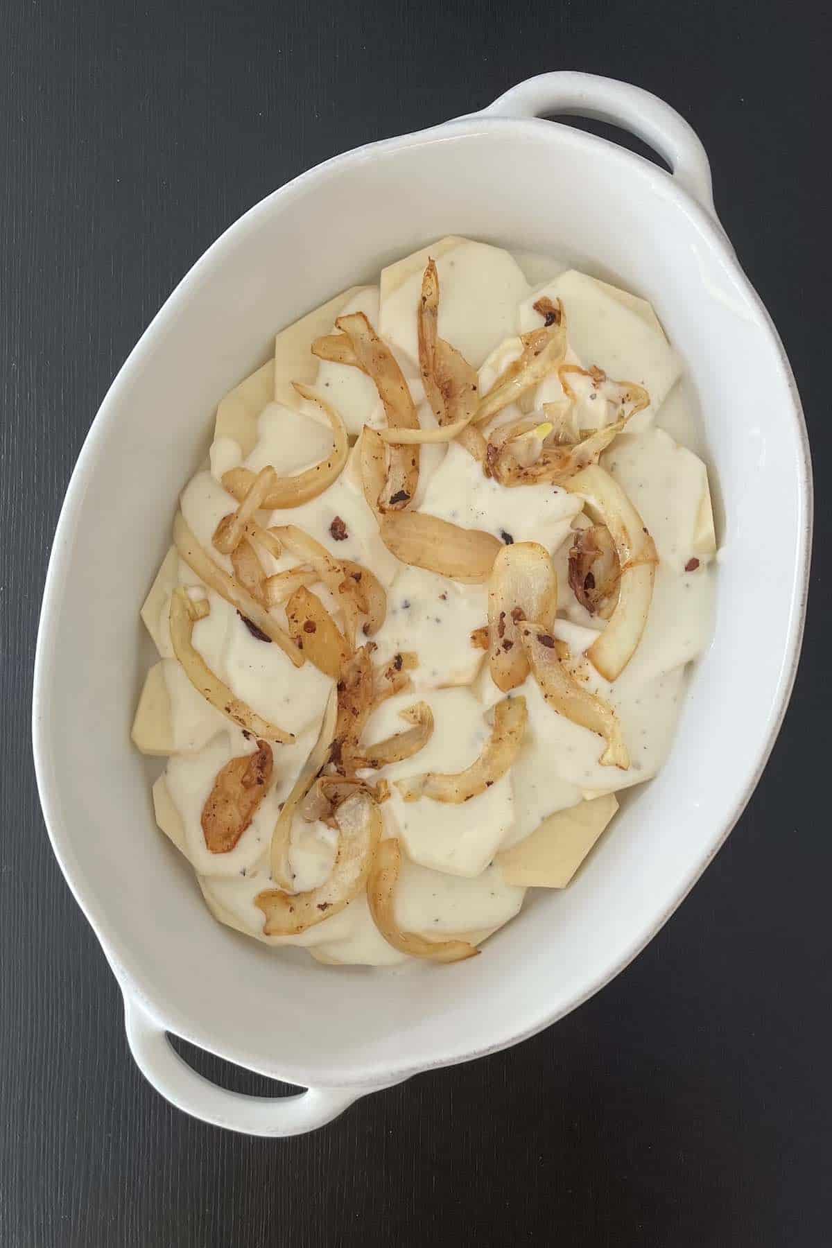 Caramelized onions distributed over the roux and potatoes in a white baking dish.