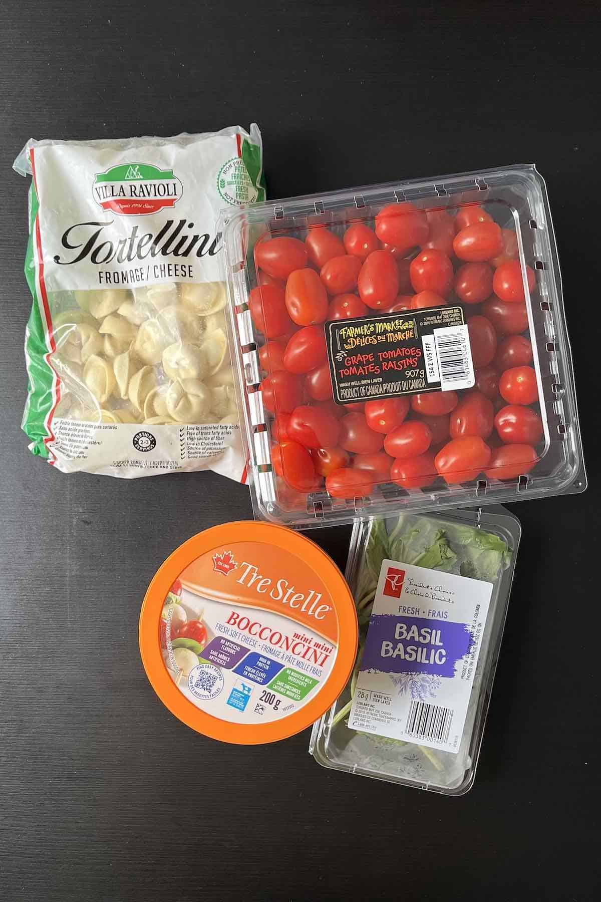 Ingredients to make the tortellini caprese skewers including cheese tortellini, cherry tomatoes, bocconcini balls, and basil on a table.