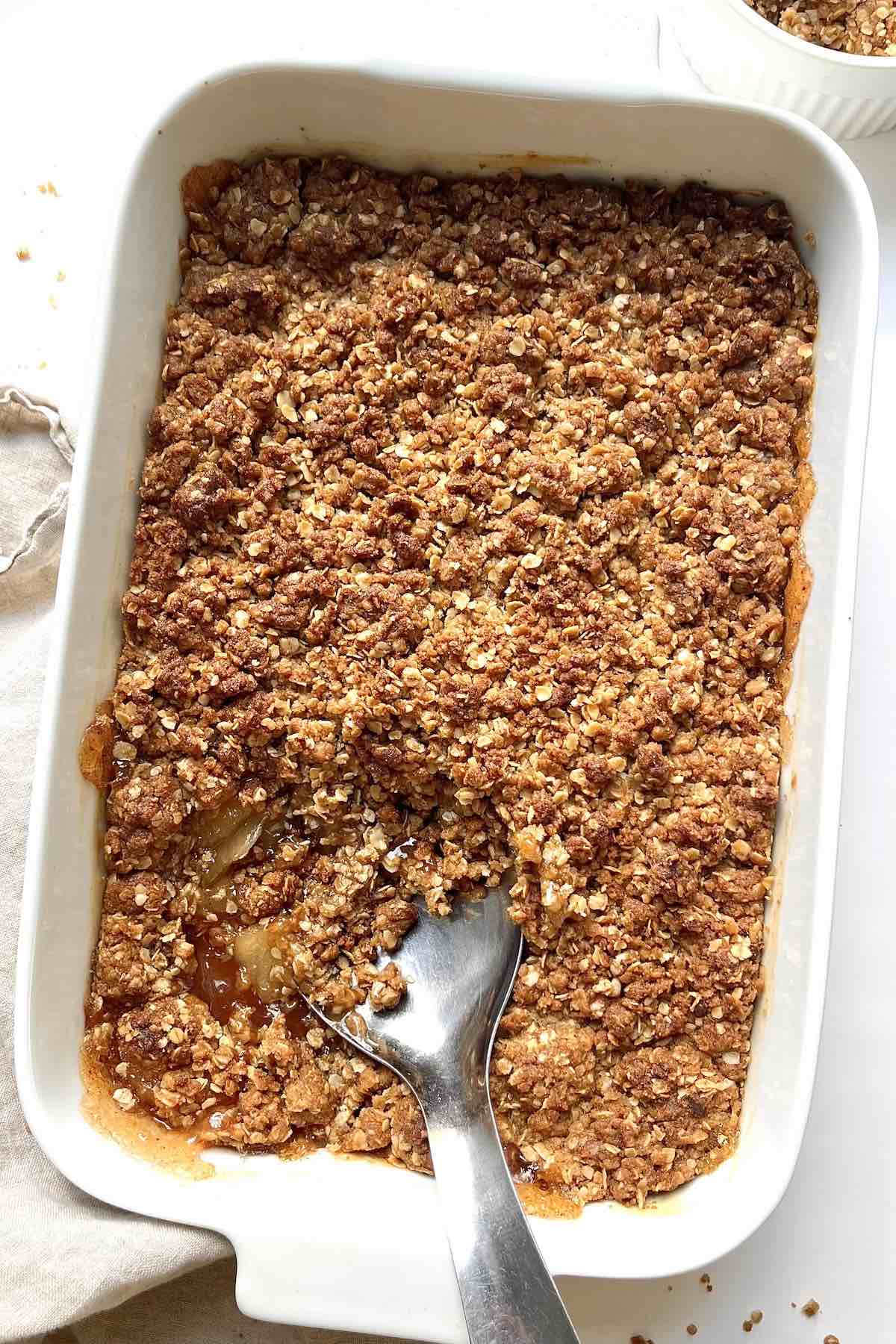 Apple crisp with canned apples in a white baking dish with a metal serving spoon in it.