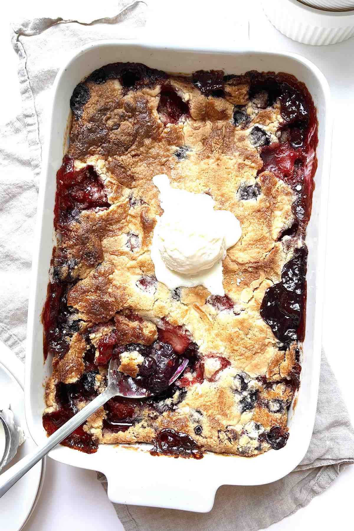 Berry cobbler in a white casserole dish topped with a scoop of vanilla ice cream.