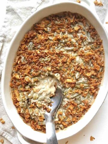 Creamy green bean casserole in a white oval casserole dish with a metal serving spoon in it.