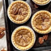 Pecan butter tarts on a baking sheet with pecans scattered around them.