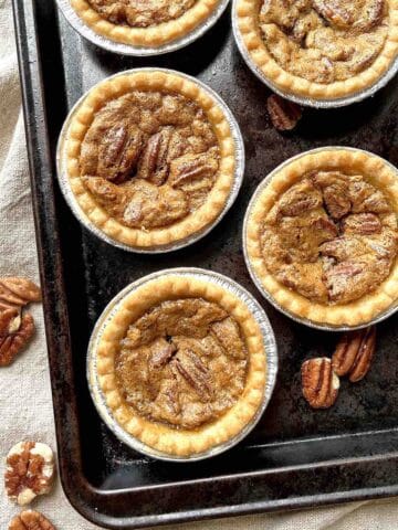 Pecan butter tarts on a baking sheet with pecans scattered around them.