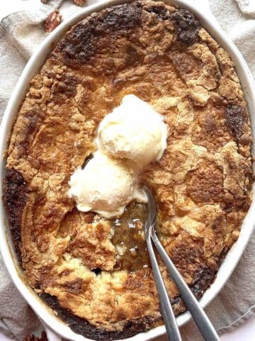 Pecan pie cobbler with cake mix in a white oval baking dish topped with two scoops of vanilla ice cream.