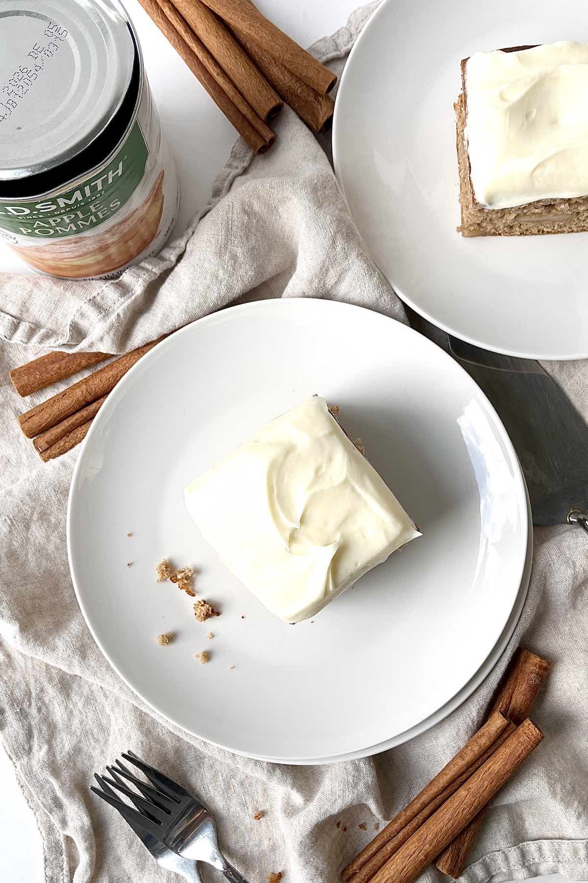 Slice of frosted apple spice cake on a white plate with cinnamon sticks and a can of pie filling next to it.