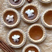 Mini pumpkin pie tarts topped with whipped cream scattered on a piece of brown parchment paper.