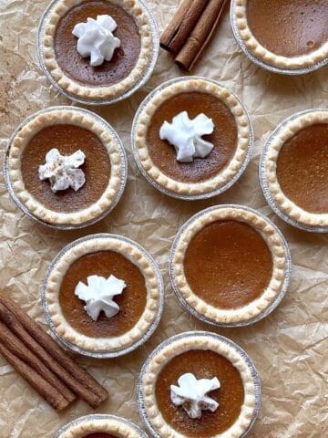 Mini pumpkin pie tarts topped with whipped cream scattered on a piece of brown parchment paper.