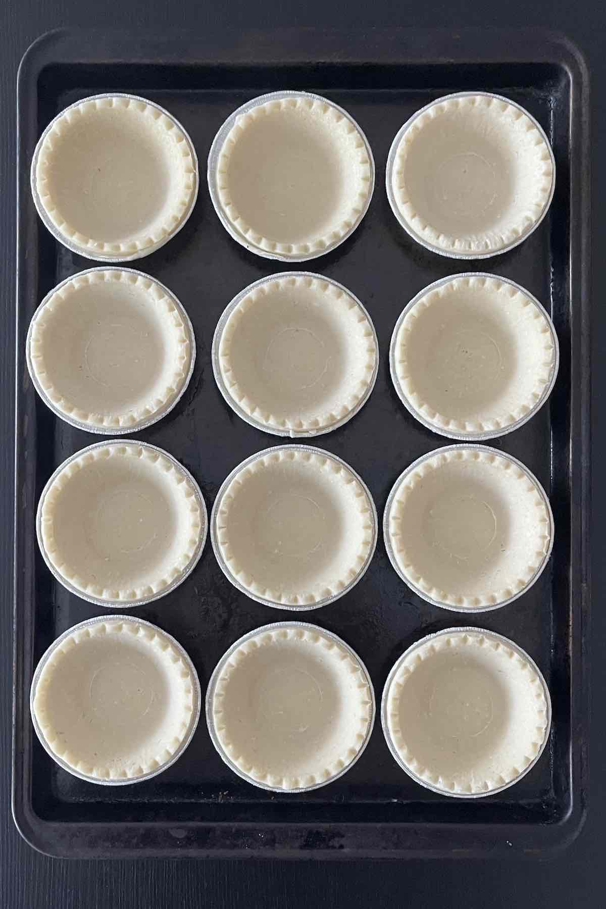 12 tart shells lined on a baking sheet.