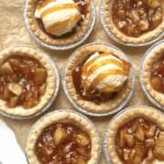 Mini apple pie tarts topped with vanilla ice cream and caramel drizzle on a piece of crinkly brown parchment paper.