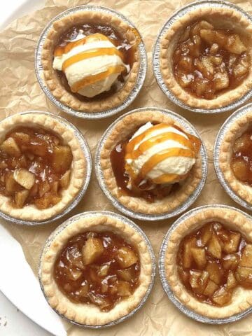Mini apple pie tarts topped with vanilla ice cream and caramel drizzle on a piece of crinkly brown parchment paper.