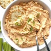 Cold buffalo chicken dip garnished with sliced green onions in a white serving bowl with two crackers and spoons in it.