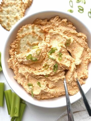 Cold buffalo chicken dip garnished with sliced green onions in a white serving bowl with two crackers and spoons in it.