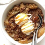 Granola apple crisp in a bowl topped with two scoops of ice cream and caramel drizzle.