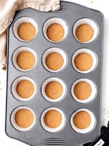 Mini graham cracker pie crusts in a muffin pan with black oven mits and a beige linen towel next to it.
